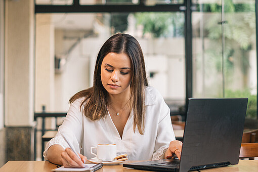 
                Eine Frau sitzt an einem Schreibtisch vor einem Laptop und macht Notizen in einem Block.
            