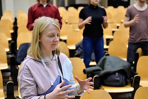 
                Studentin mit drei Studierenden im Hintergrund bei einem Workshop der Kompetenz-Werkstatt in einem Hörsaal.
            