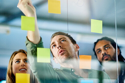 Psychologische Grundlagen der Personalführung in der Öffentlichen Verwaltung
Drei Personen beschreiben eine Glaswand, an der Klebezettel in verschiedenen Farben angebracht sind.